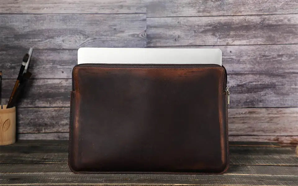 Brown leather laptop sleeve with a laptop inside on a wooden surface.