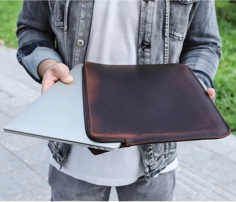 Person holding a brown leather laptop sleeve over a silver laptop outdoors.